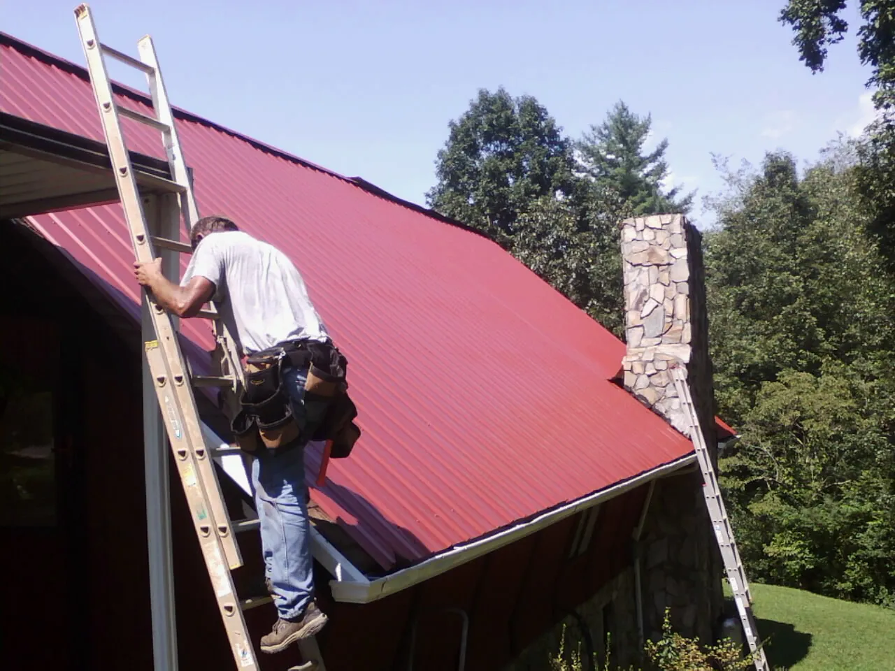 Roofer on a ladder beside a red metal roof and stone chimney — Soffit & Fascia Repair by Roofing Brothers in Mullica Hill
